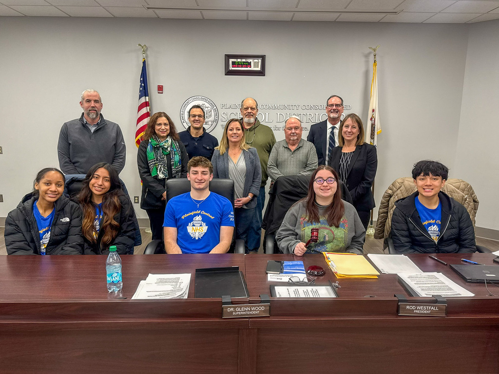 group of students sit at board table. Board members and superintendent of schools stand behind them