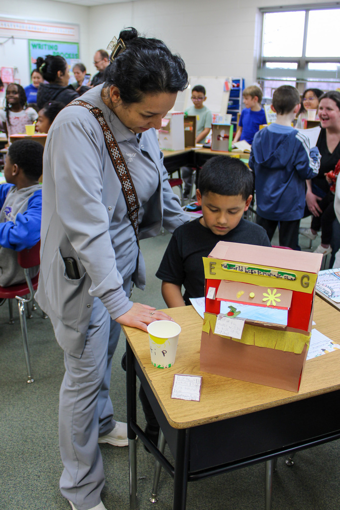 Woman stands next to seated male child who has a homemade lemonade shoebox stand on desk