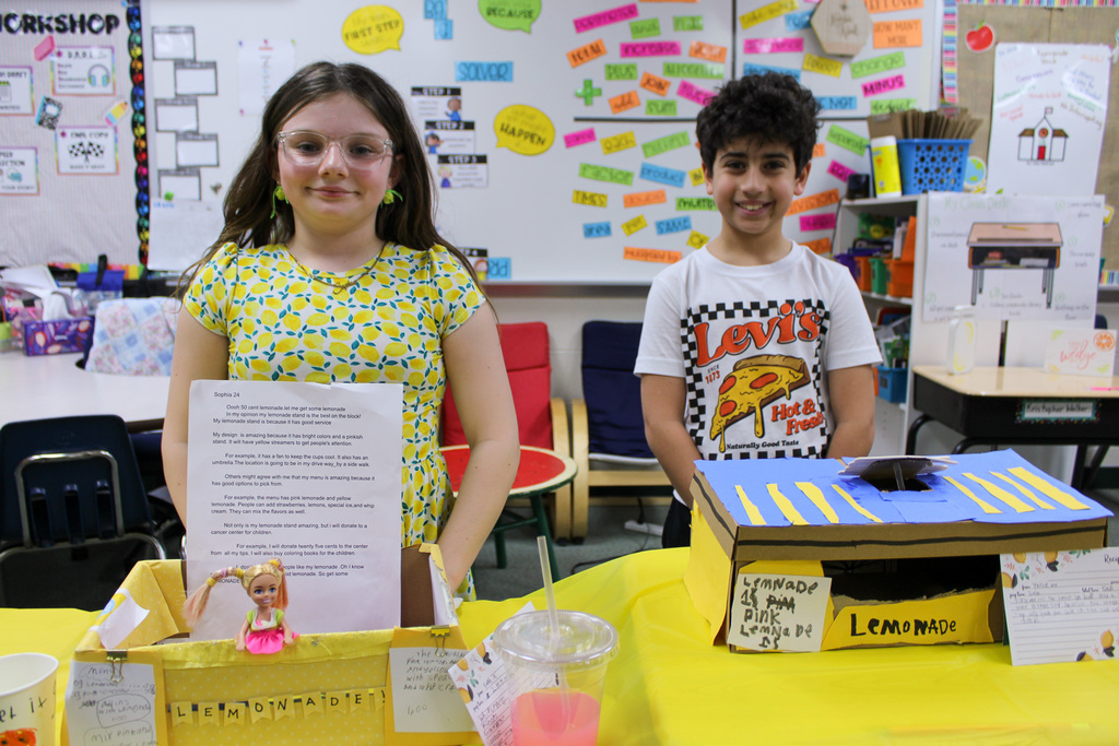 boy and girl stand behind shoebox lemonade stands in classroom