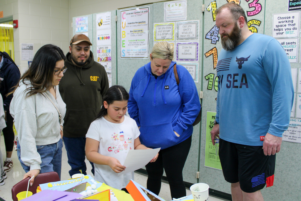 group of adults stand around girl who is reading paper to them