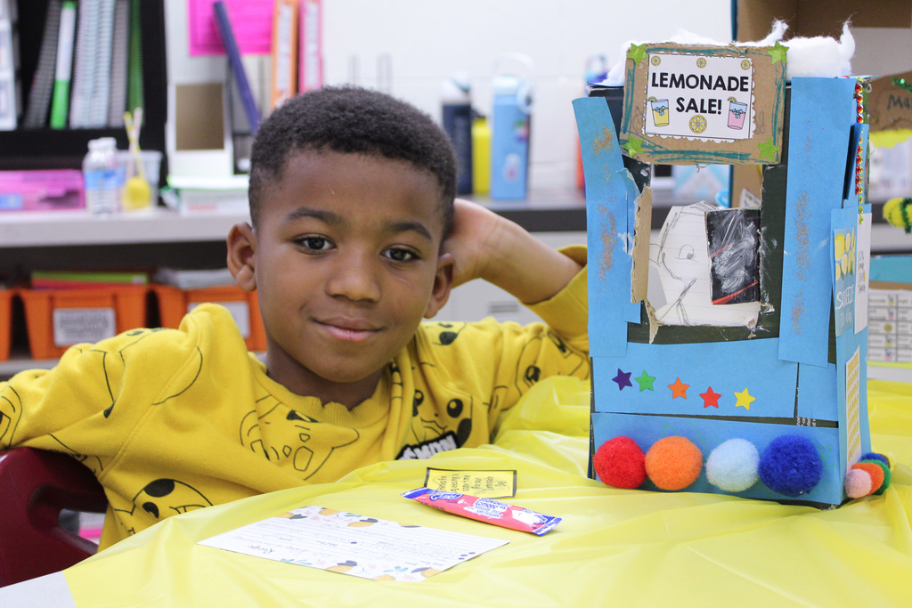 boy wearing yellow shirt sits next to blue shoebox decorated like a lemonade stand