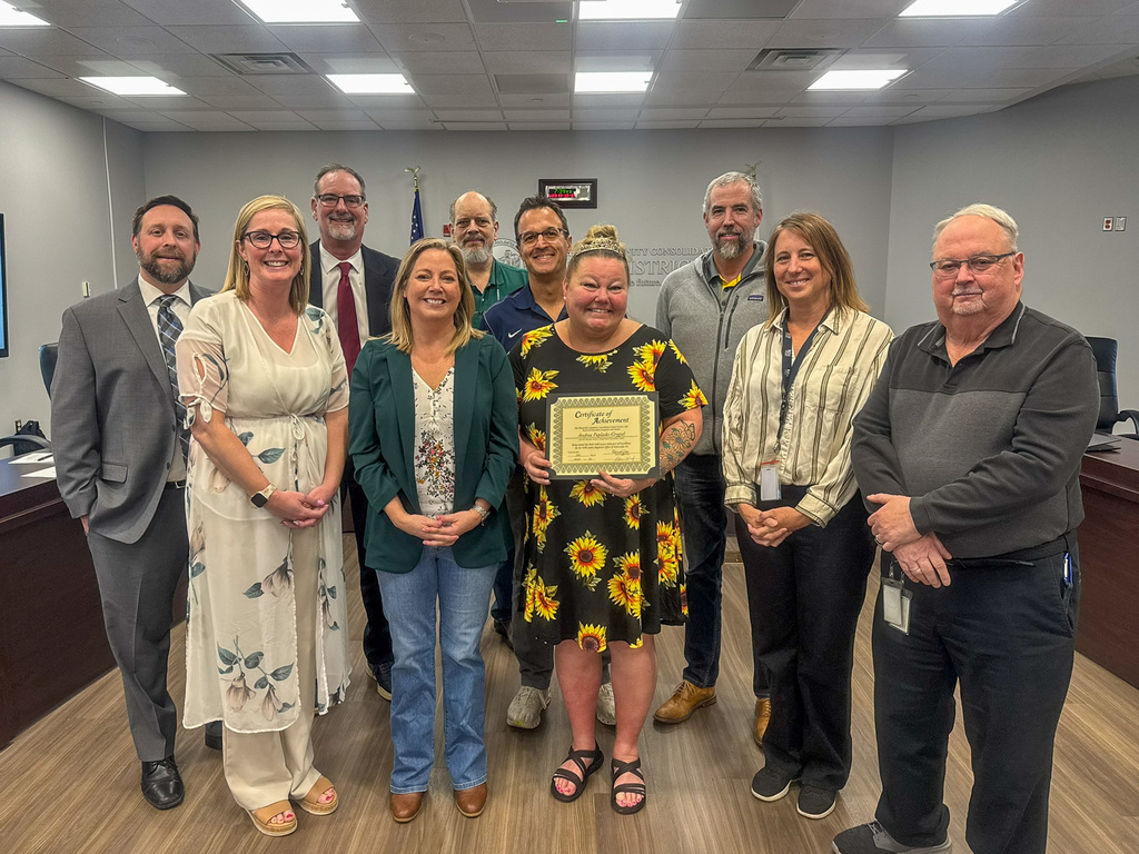 group of men and woman standing in board room around woman in black sunflower dress holding certificate