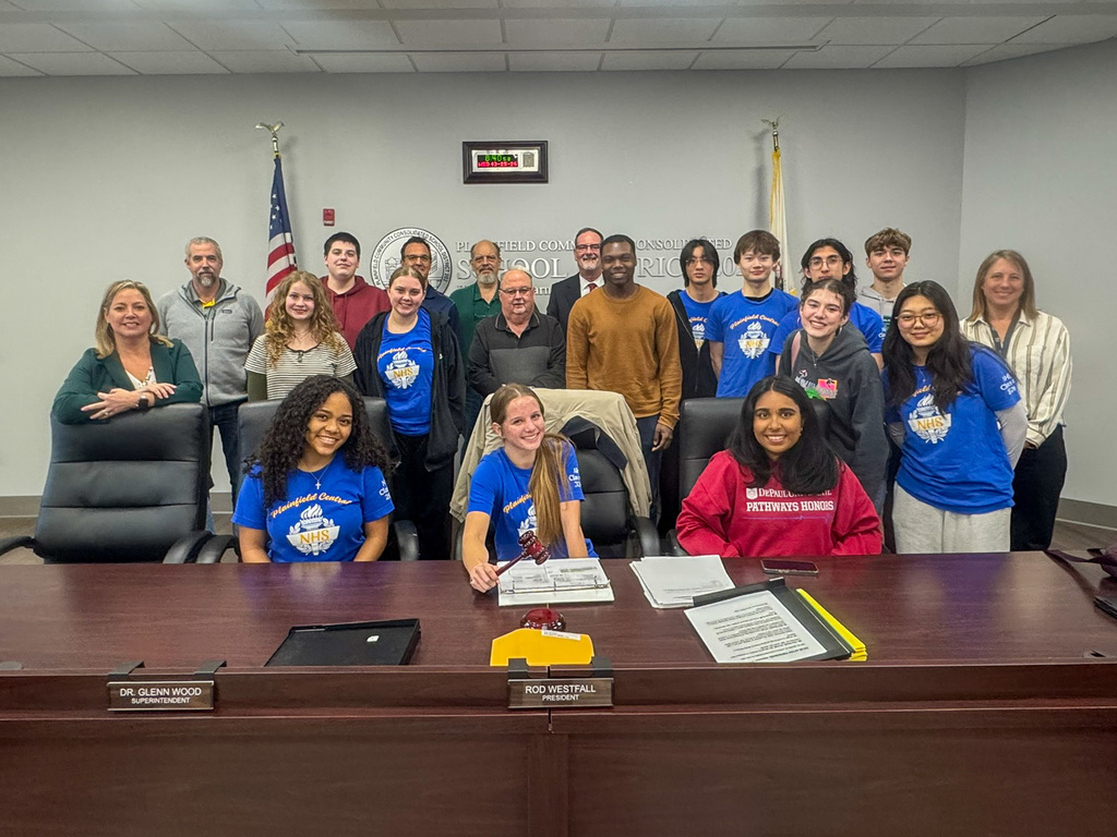 group of students stand and sit at desk with Board of Education members