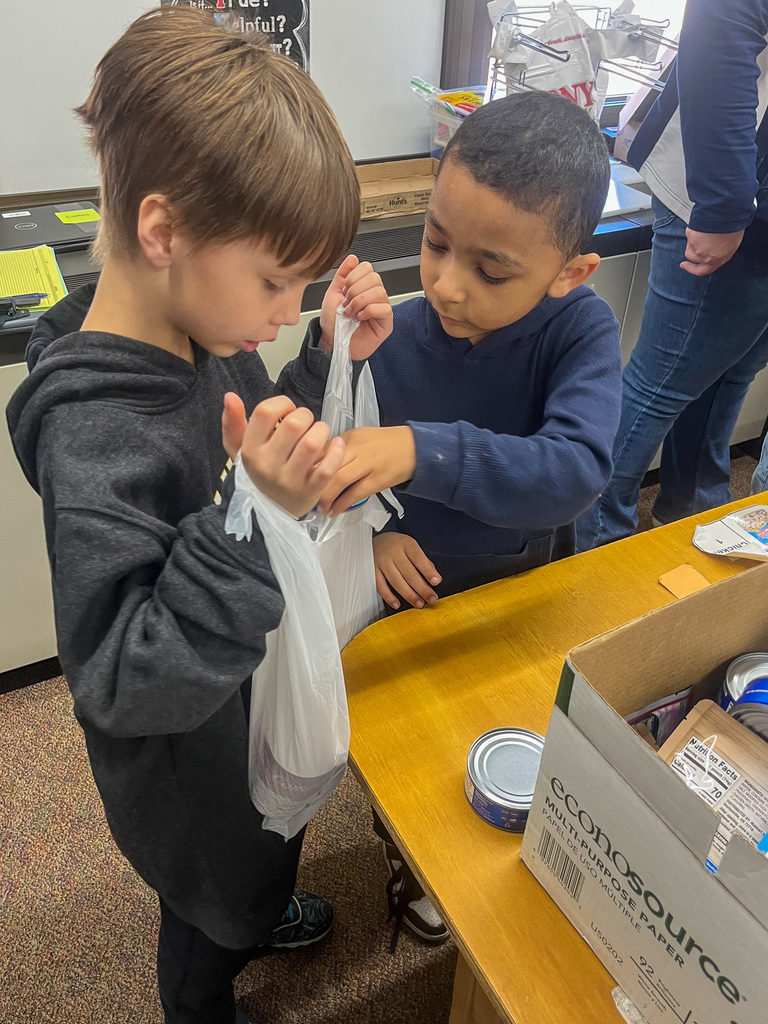 two elementary school male students put canned goods into a plastic bag