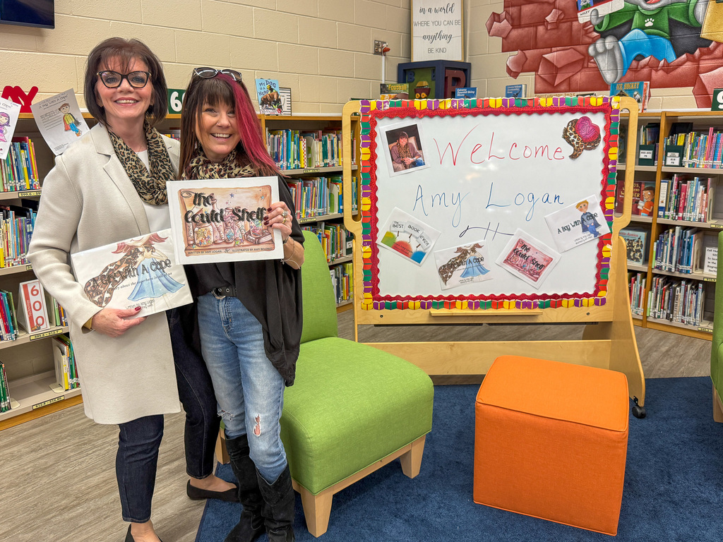 Two adults stand together holding books. White board reads Welcome Amy Logan.