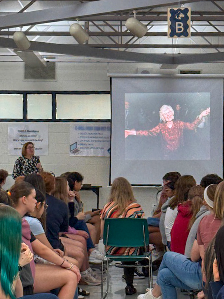 group of students sit listening to speaker and look at screen with an older woman with white hair dancing