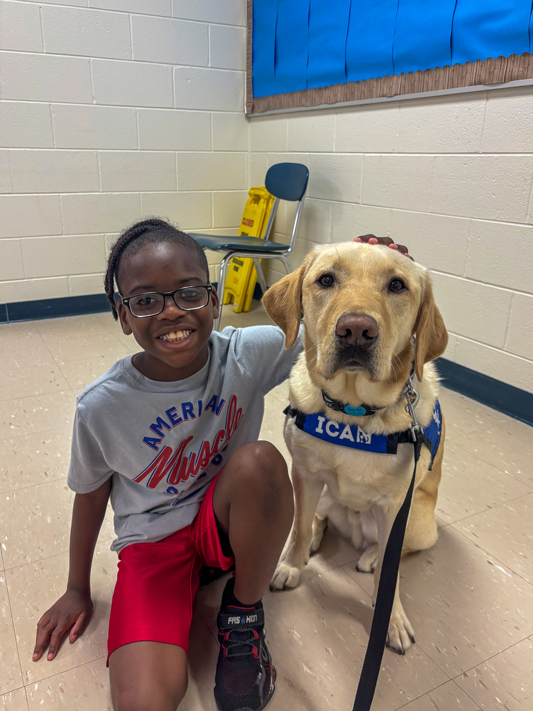 an elementary school male student sits on floor with arm around Facility Dog Tater Tot