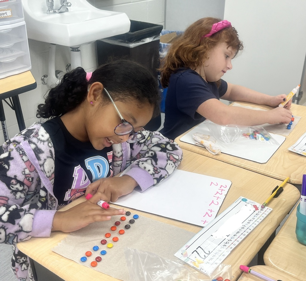 Two students sitting in a classroom, counting M&Ms and writing numbers on a whiteboard.