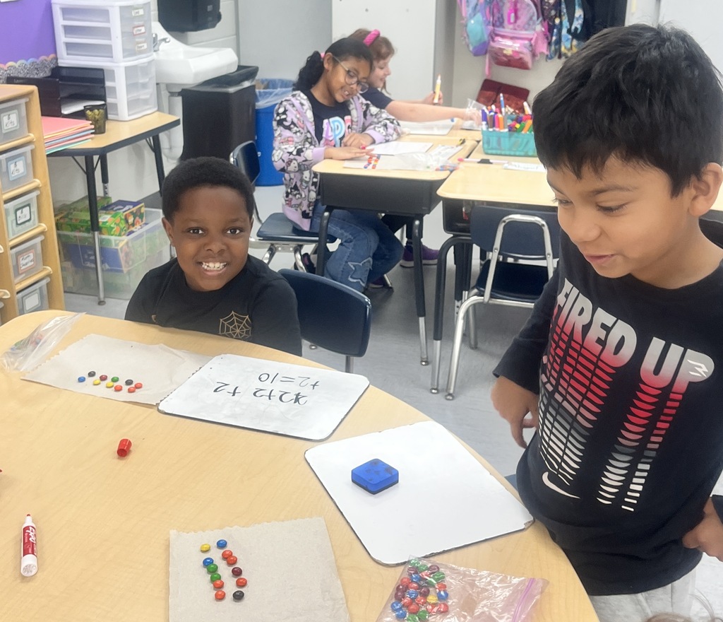 Two students sitting in a classroom, counting M&Ms and writing numbers on a whiteboard.