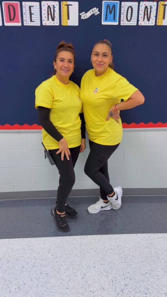 Two teachers in yellow shirts and black pants smiling in the hallway.