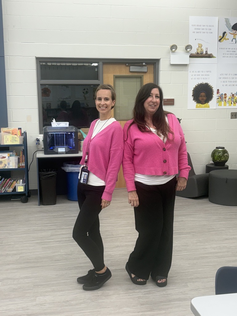 Two teachers wearing pink sweaters and black pants standing in a library.