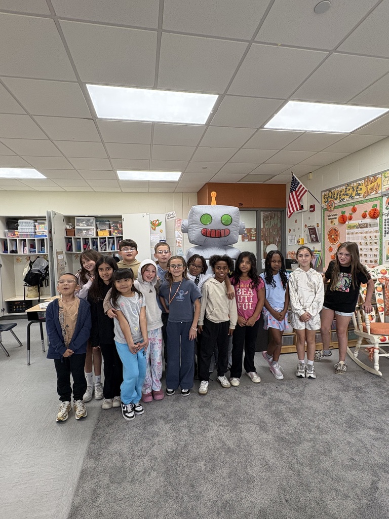 Students standing in a classroom with a teacher, dressed as a robot.