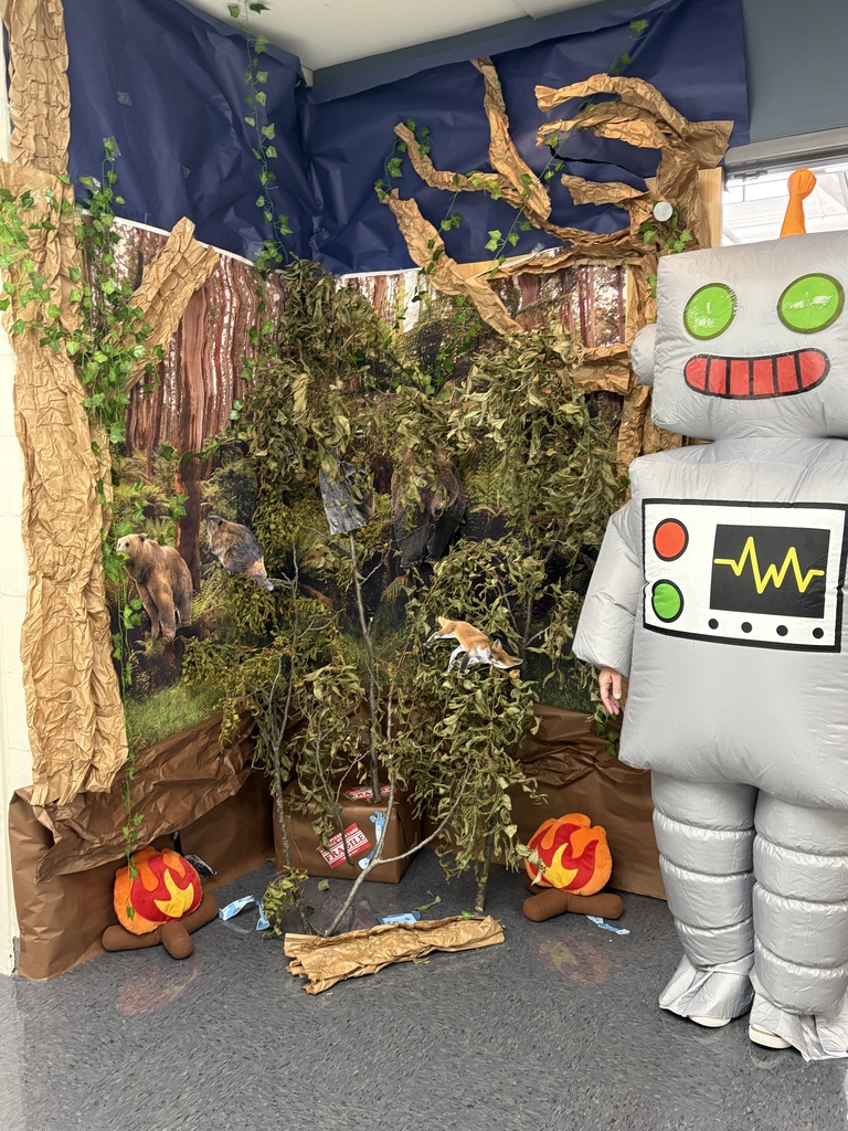 A teacher in a robot costume standing next to a hallway display with a forest, backdrop and fake campfires.