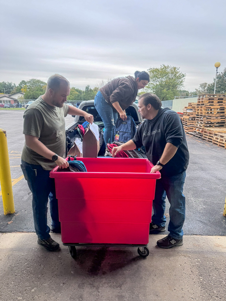 three people unloading backpacks from the back of a truck into a red bin