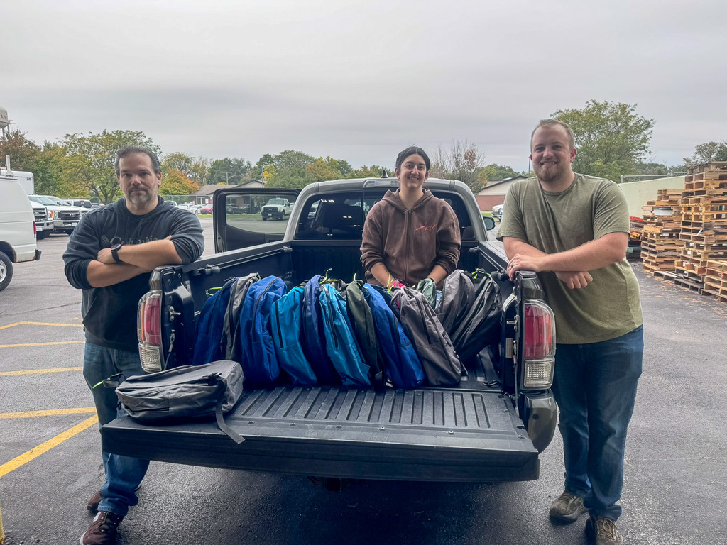 Three people pose in the back and sides of a truck with box and backpack donations