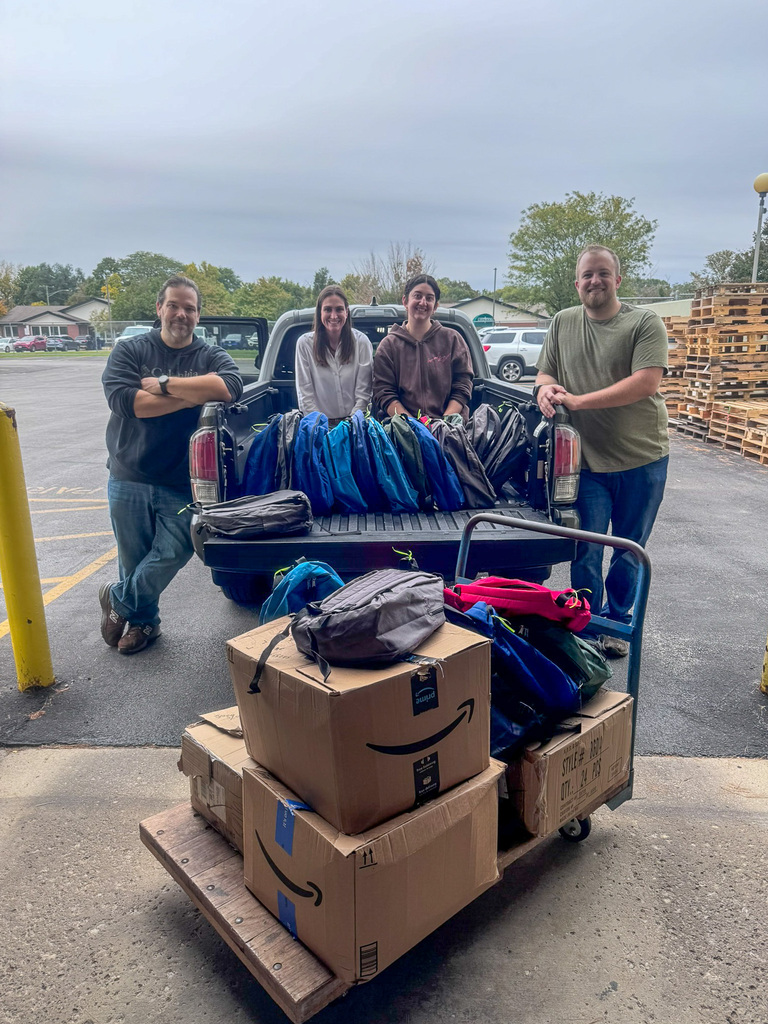 Four people pose in the back and sides of a truck with box and backpack donations