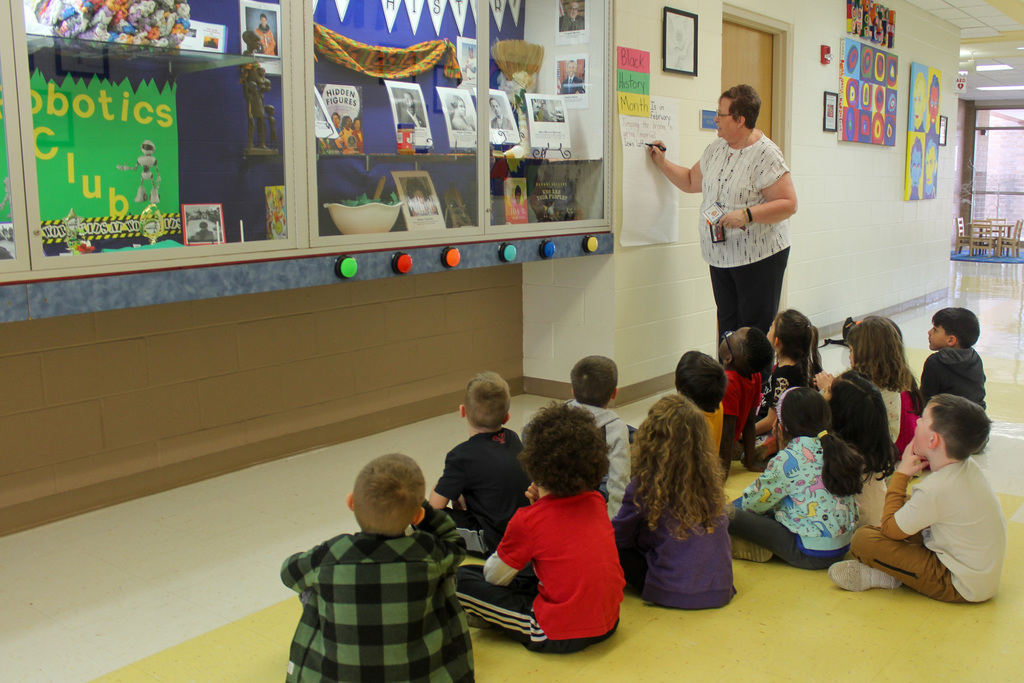 kindergarten students sit in front of a display case while a teacher writes about Black History Month on a poster