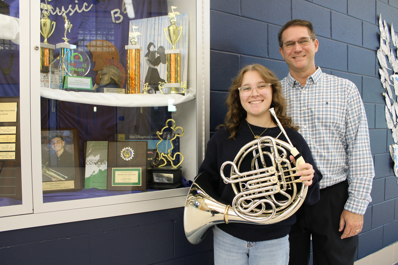 student holds French horn, man stands behind her near trophy case