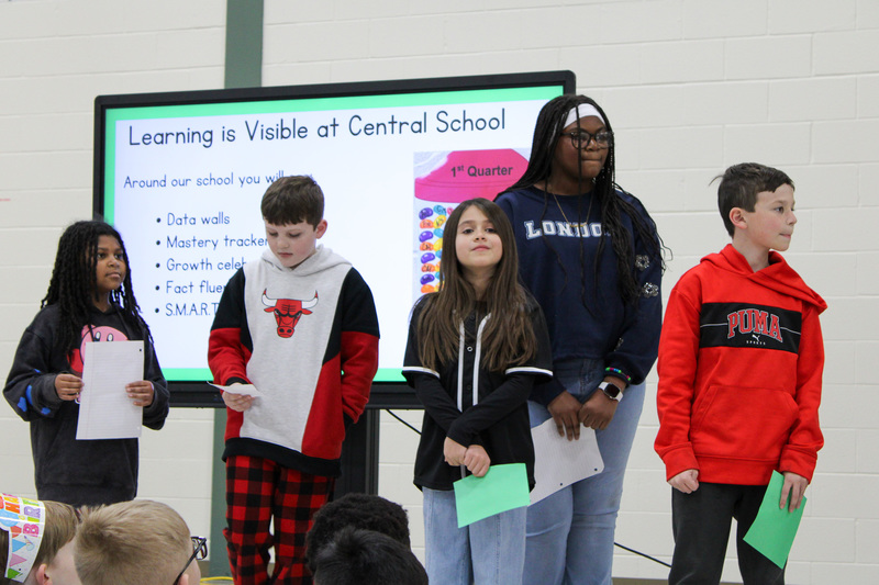 five students holding papers stand in front of large screen