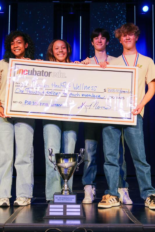 four students stand on stage holding oversize check in front of a trophy
