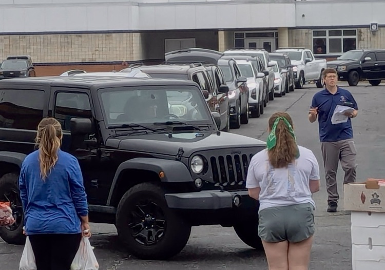 picture of three staff members handing out food service bags to a line of cars outside school building