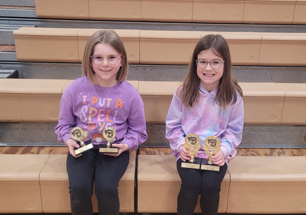 two young ladies who won the local spelling bee sitting on the bleachers with their trophies
