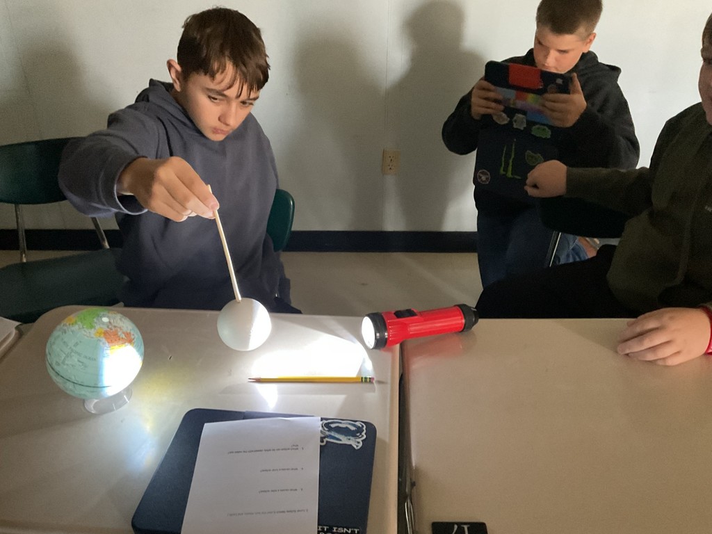Sixth grade science students use globes, styrofoam balls, and flashlights to model solar and lunar eclipses, demonstrating how the Sun, Earth, and Moon align to create shadows during an eclipse.