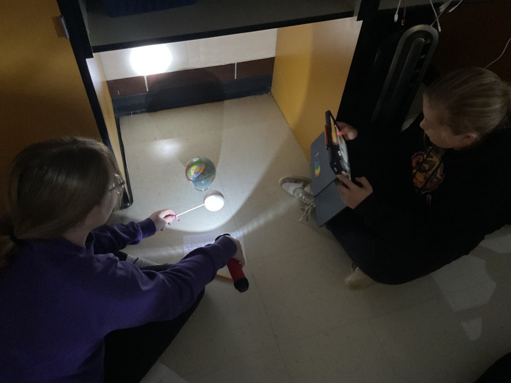 Sixth grade science students use globes, styrofoam balls, and flashlights to model solar and lunar eclipses, demonstrating how the Sun, Earth, and Moon align to create shadows during an eclipse.