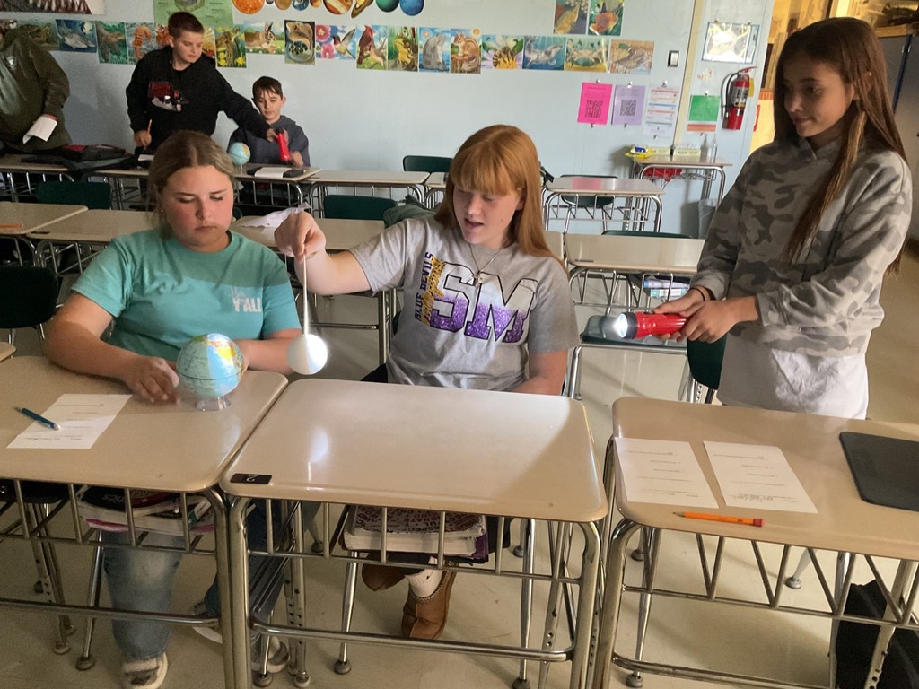 Sixth grade science students use globes, styrofoam balls, and flashlights to model solar and lunar eclipses, demonstrating how the Sun, Earth, and Moon align to create shadows during an eclipse.