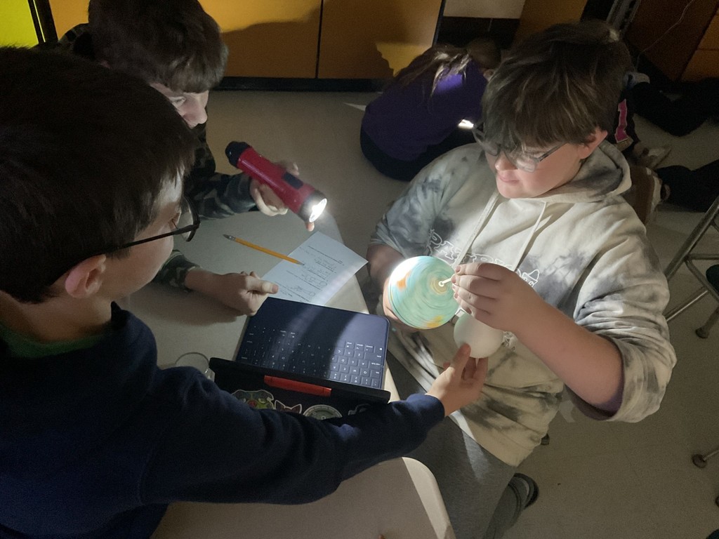 Sixth grade science students use globes, styrofoam balls, and flashlights to model solar and lunar eclipses, demonstrating how the Sun, Earth, and Moon align to create shadows during an eclipse.