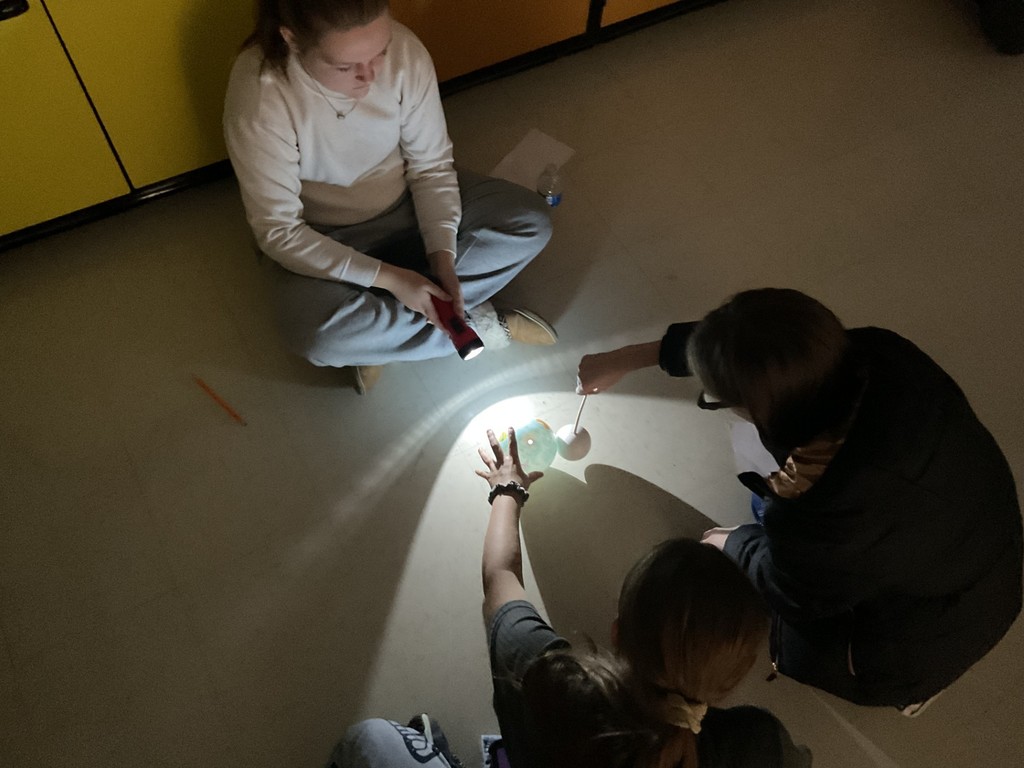 Sixth grade science students use globes, styrofoam balls, and flashlights to model solar and lunar eclipses, demonstrating how the Sun, Earth, and Moon align to create shadows during an eclipse.