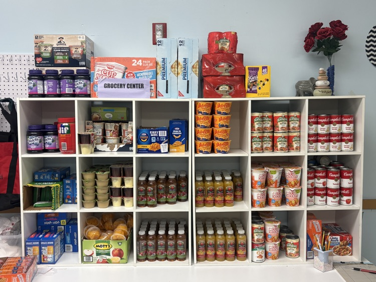 White cubby shelves filled with assorted pantry items such as canned goods, noodles, snacks, applesauce cups, and juice bottles, with a “Grocery Center” sign displayed in the middle.