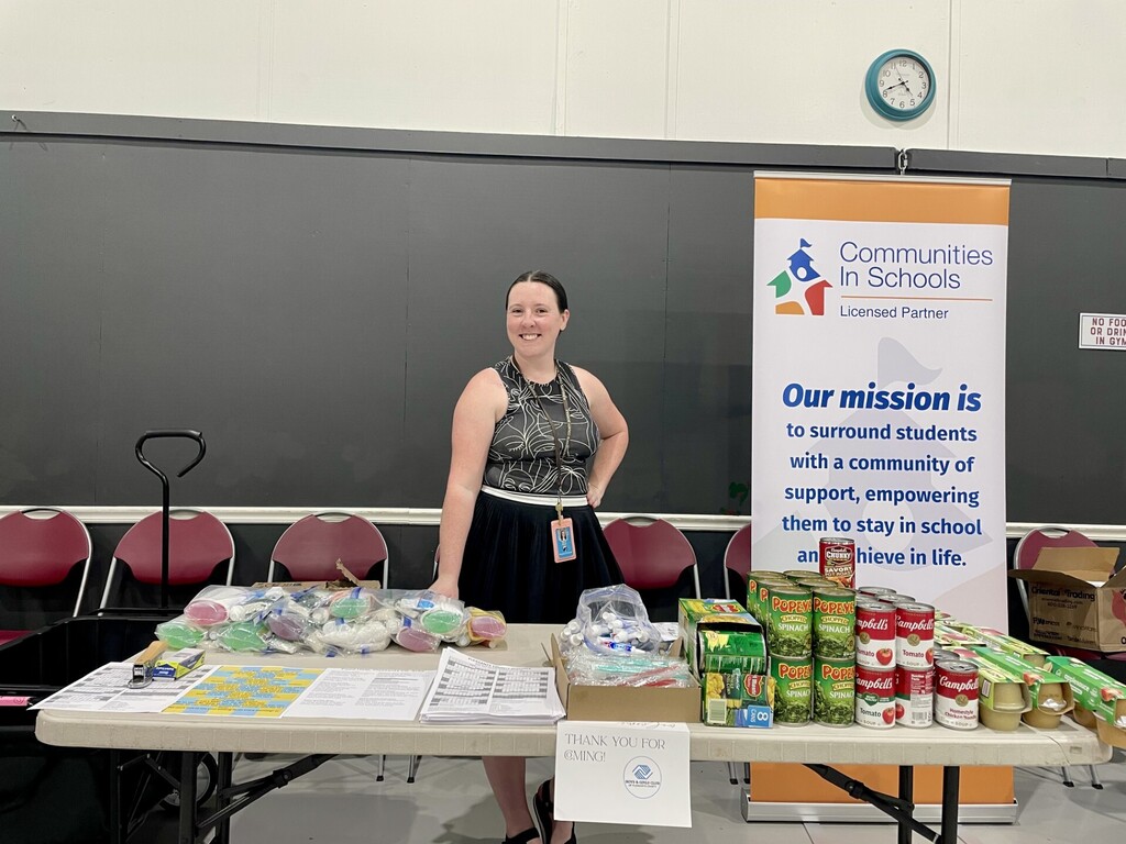 A smiling woman stands behind a table at the Pleasants County Back to School Fair, representing Belmont Elementary School and Communities In Schools. The table displays family resources, personal care items, school supplies, and donated canned and packaged foods. A Communities In Schools banner behind her reads, “Our mission is to surround students with a community of support, empowering them to stay in school and achieve in life.”