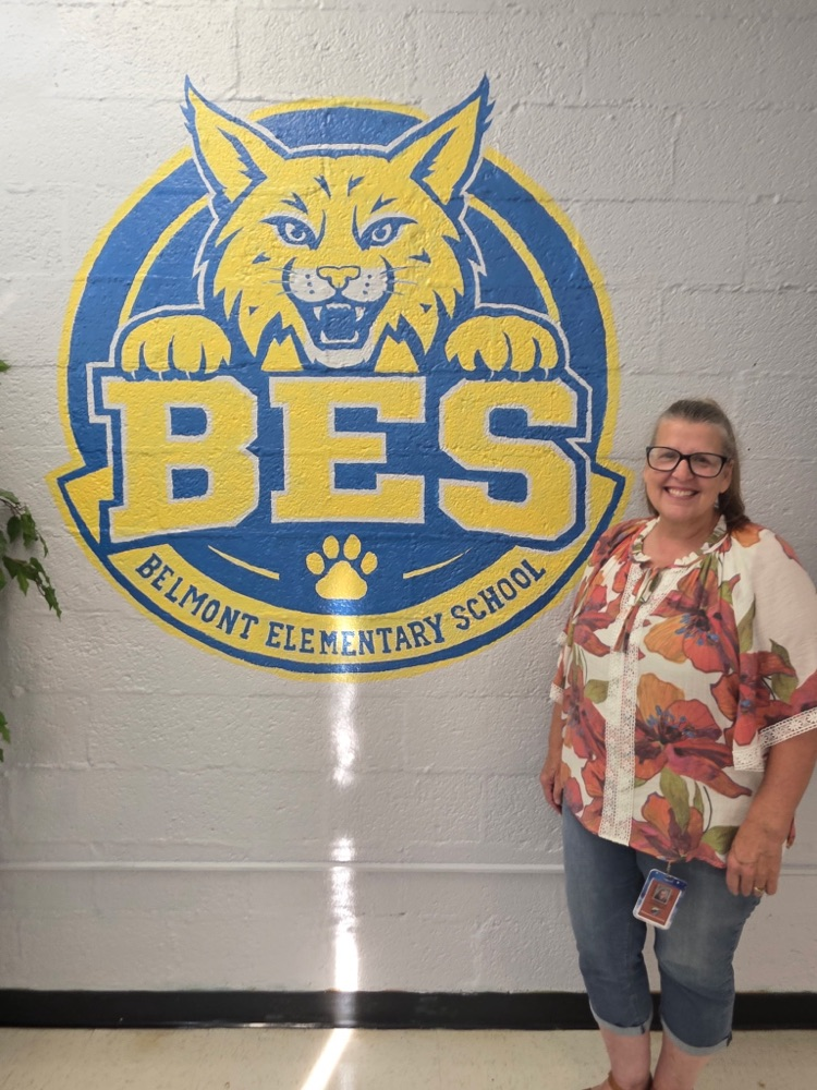 Mrs. VanValey stands smiling beside a colorful hand-painted mural in the new Belmont Elementary School conference room. The mural features a large blue and gold Bobcat logo with the letters “BES” and “Belmont Elementary School” underneath.