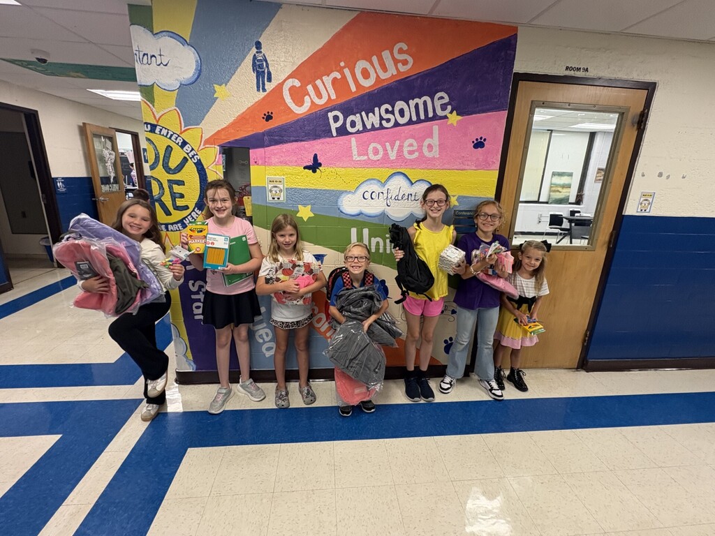Several elementary students stand in a school hallway holding donated backpacks and school supplies. A table nearby displays additional backpacks, crayons, pencils, notebooks, folders, scissors, and other classroom supplies. The donation was generously provided by Matheny Ford and the Pleasants County Chamber of Commerce.