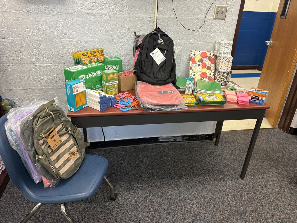 Several elementary students stand in a school hallway holding donated backpacks and school supplies. A table nearby displays additional backpacks, crayons, pencils, notebooks, folders, scissors, and other classroom supplies. The donation was generously provided by Matheny Ford and the Pleasants County Chamber of Commerce.
