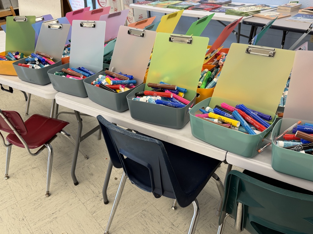 A row of colorful instructional gift baskets sits on classroom tables, each filled with markers and supplies and topped with pastel clipboards.