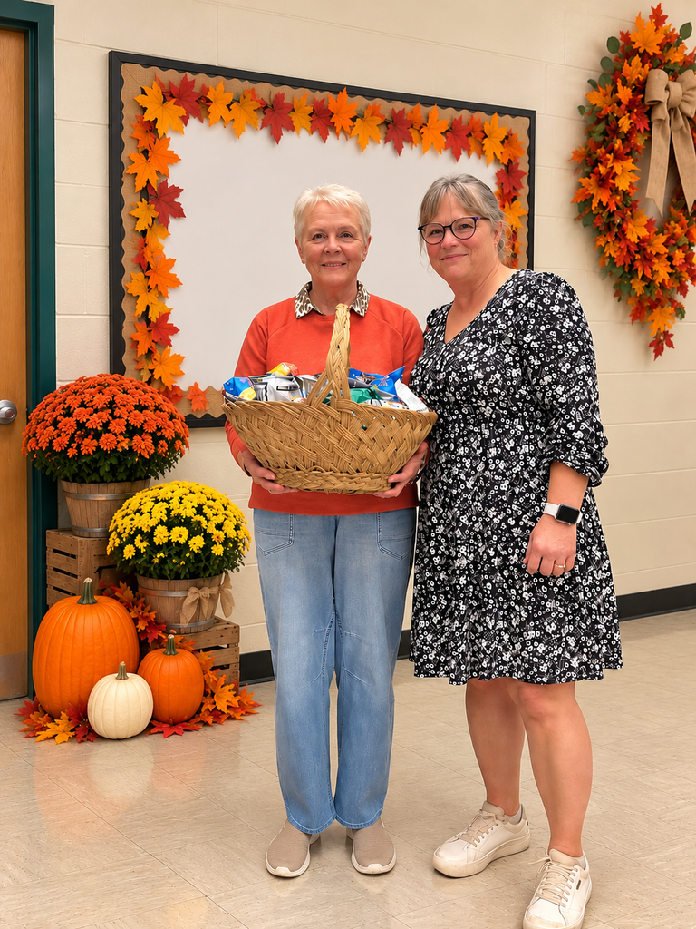 A fall-themed appreciation basket is presented in a warmly decorated school hallway, surrounded by colorful autumn leaves, pumpkins, and potted mums. The seasonal display creates a welcoming atmosphere as two individuals pose with the gift basket, celebrating gratitude, recognition, and community spirit.