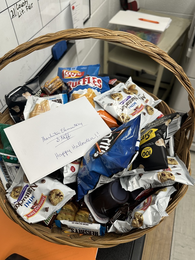 A fall-themed appreciation basket is presented in a warmly decorated school hallway, surrounded by colorful autumn leaves, pumpkins, and potted mums. The seasonal display creates a welcoming atmosphere as two individuals pose with the gift basket, celebrating gratitude, recognition, and community spirit.