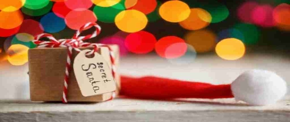 small package tied with a ribbon and tag that says "Secret Santa" on it, with the top of a Santa hat visible behind it and blurry holiday lights in the background