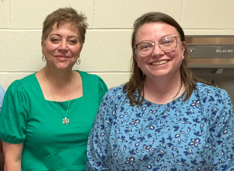 Two staff members stand side by side in front of a light-colored cinderblock wall. The person on the left wears a solid green top and necklace, and the person on the right wears a blue patterned top.