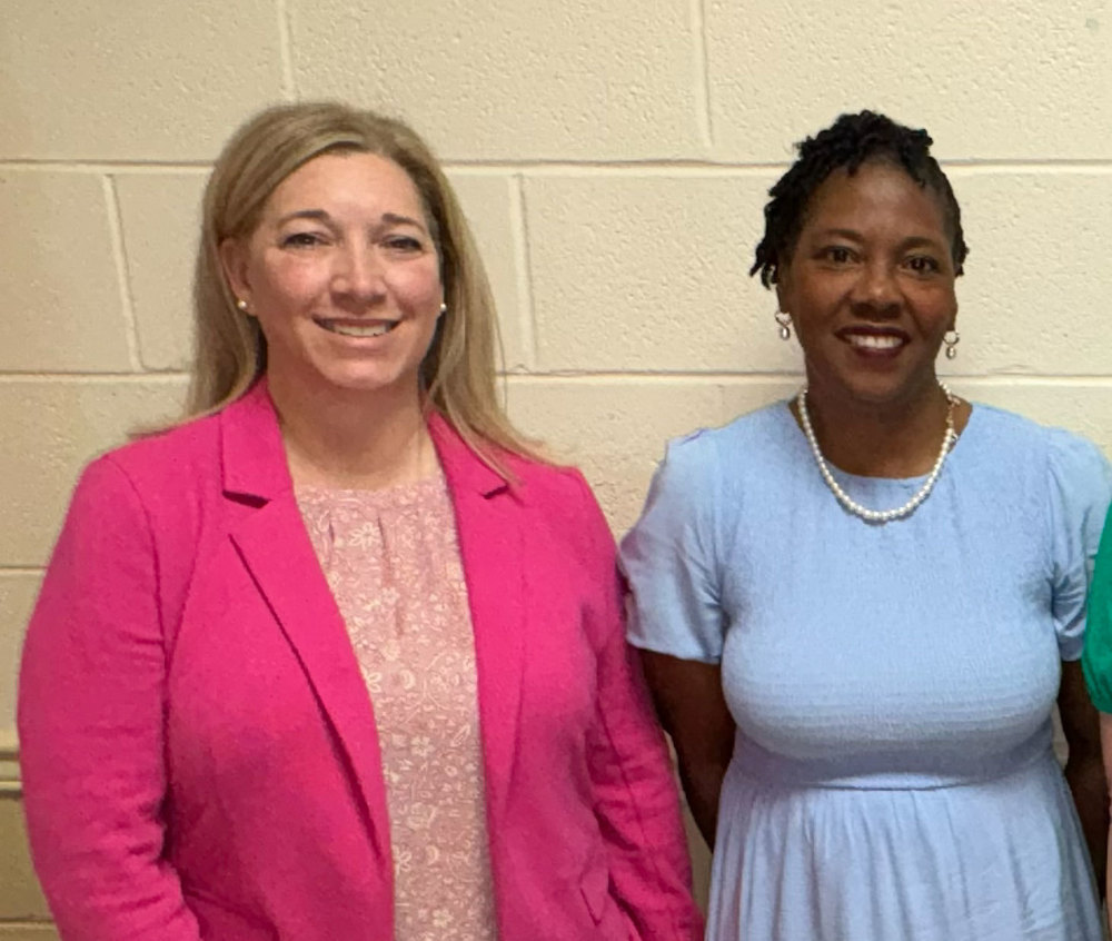 Two women stand side by side in front of a light-colored cinderblock wall. The woman on the left wears a bright pink blazer over a link pink patterned blouse, and the woman on the right wears a light blue short-sleeved dress with a pearl necklace and earrings. Both women are shown from aboujt mid-torso up, facing the camera.