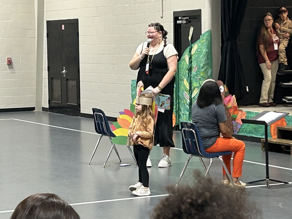 Students in thanksgiving costumes on a stage. Teacher speaking into microphone