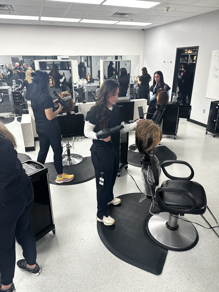Cosmetology students working at stations holding hair dryers and working mannequins.