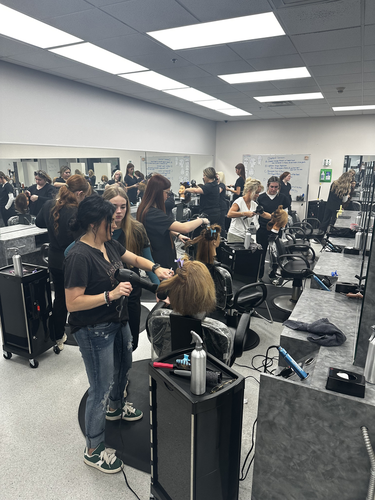 Cosmetology students working at stations holding hair dryers and working mannequins.