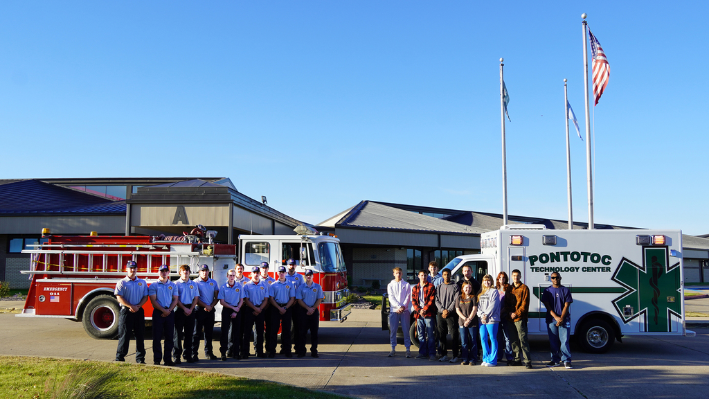 Two groups of students standing in front of a fire engine and an ambulance against a blue sky with flags on poles in the background