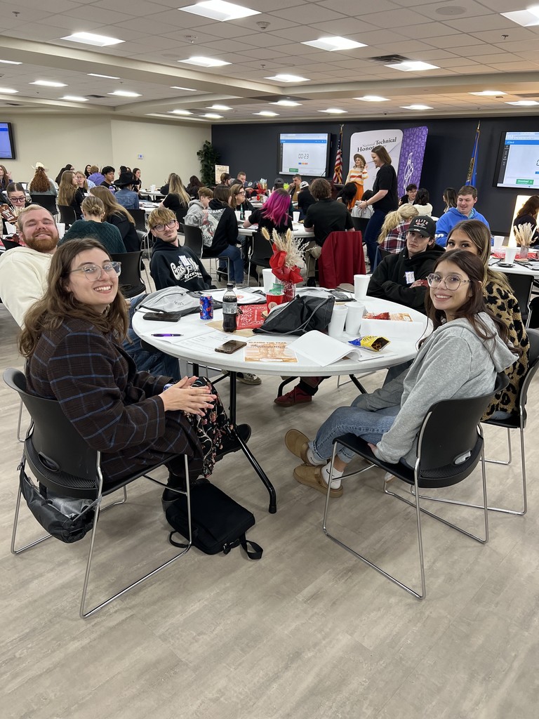 People sitting around a round table and chairs with lots of other people and tables in the background