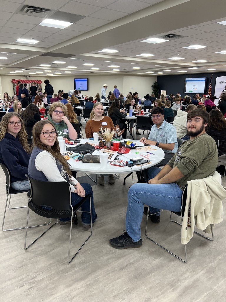 People sitting around a round table and chairs with lots of other people and tables in the background
