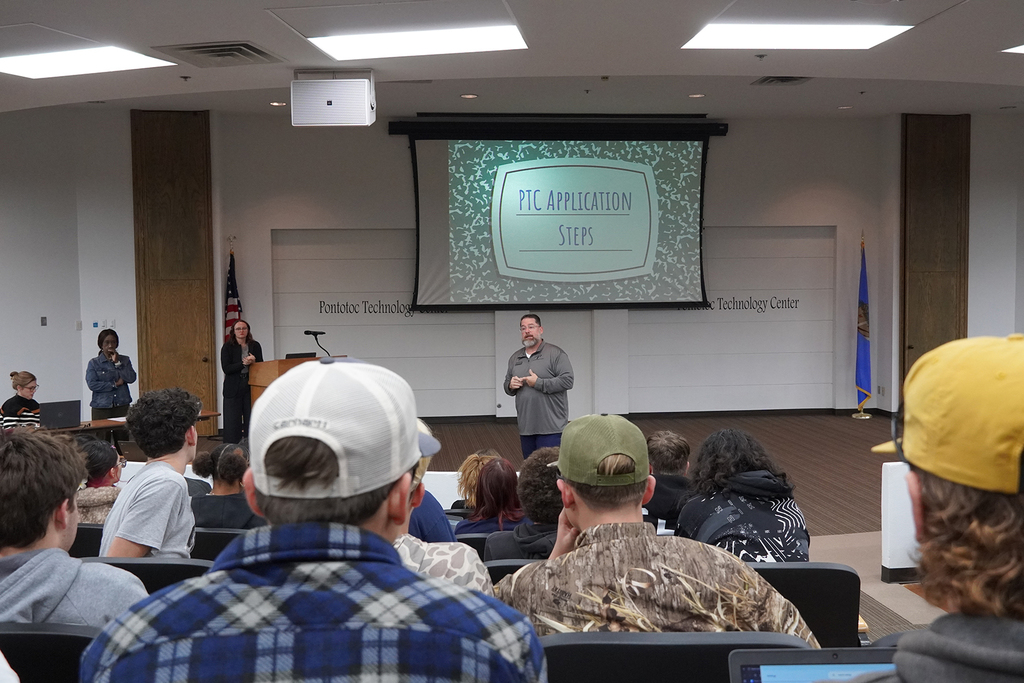 A man talking to a crowd of students in an auditorium with a projector screen behind him.