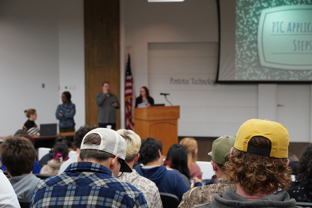 A group of students sitting in an auditorium with presenters preparing to speak at a podium.